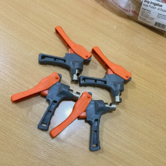 Four handheld drip irrigation hole punch tools with bright orange spring-loaded handles, dark grey ergonomic bodies, and metallic steel cutting tip nozzles arranged on a light wooden countertop